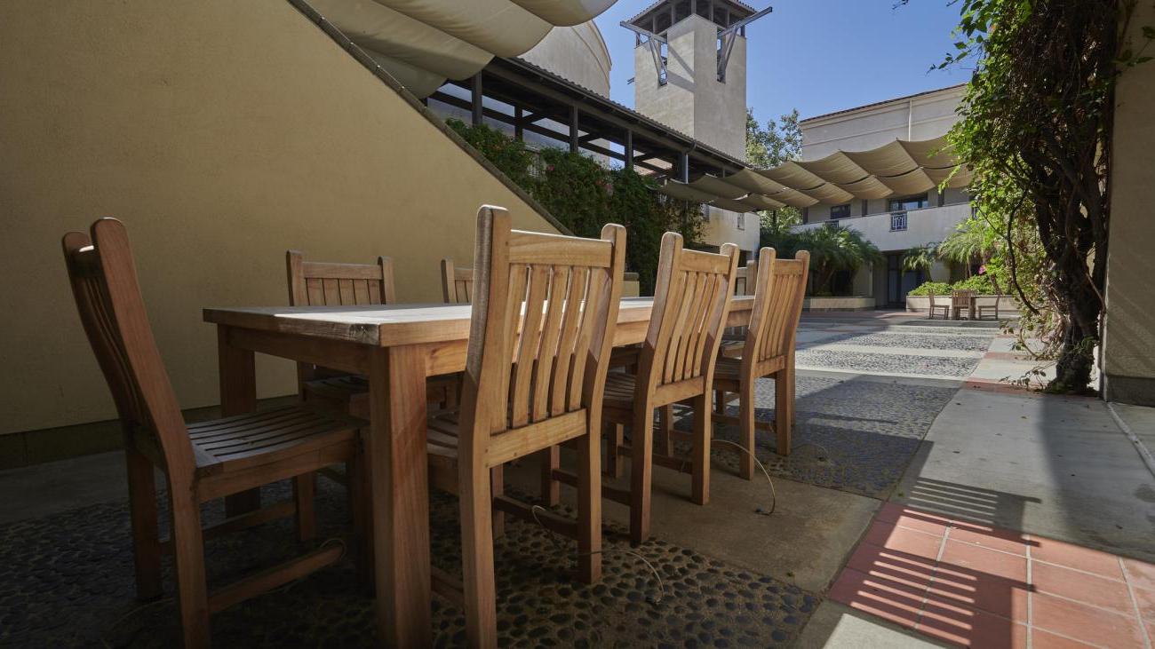 an outdoor space with a wooden chair with  8 chairs and with greenery around with stairs leading up to the second floor.